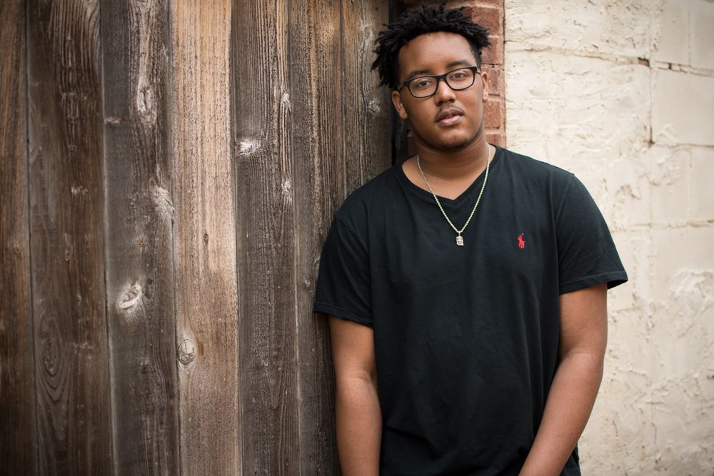A person wearing glasses and a black polo shirt stands against a weathered wooden and concrete wall.