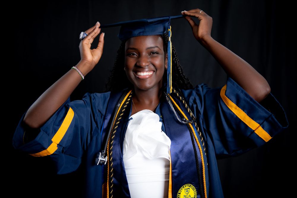 Graduate in navy blue cap and gown with gold honors cords smiles while adjusting mortarboard during graduation photo.