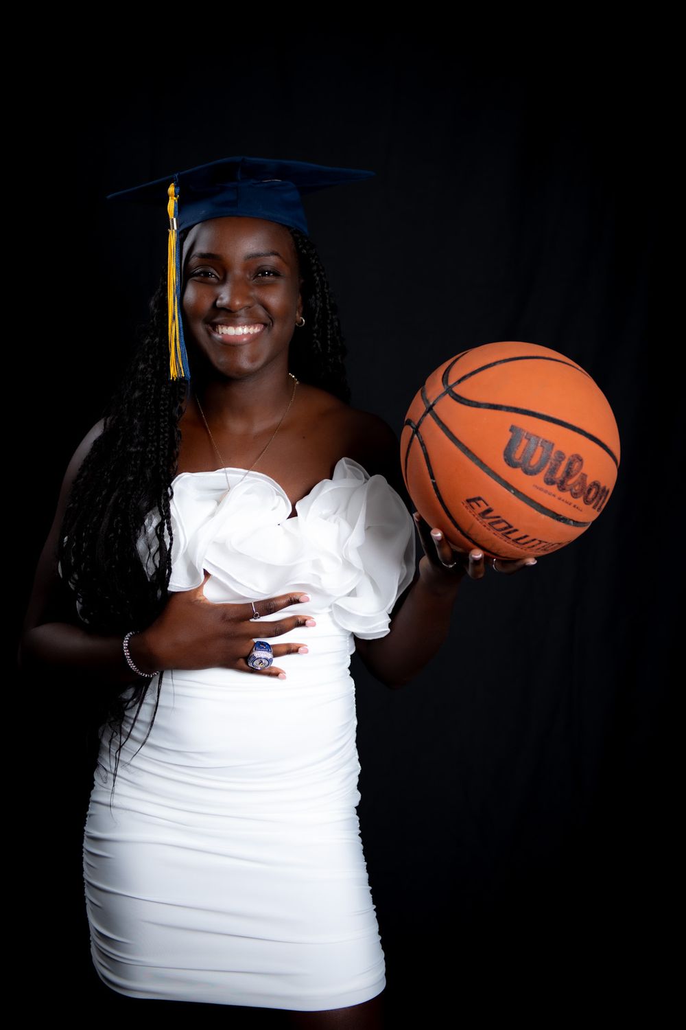 Graduate in cap and gown poses in white dress with basketball against dark background for senior photos.