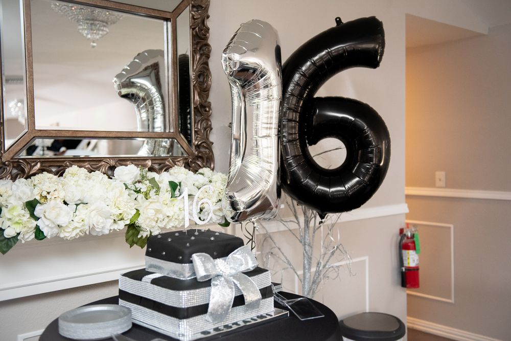 Black and silver number 16 balloons with white floral decorations and wrapped gifts on display table.