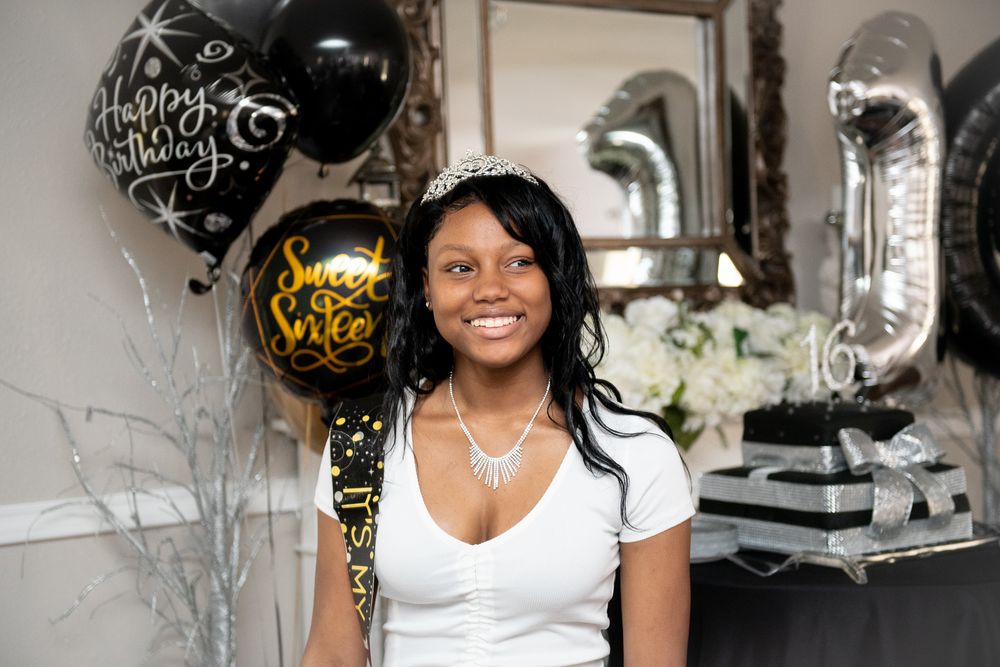 A person in a white top smiles while posing with black and metallic birthday balloons in an elegant setting.