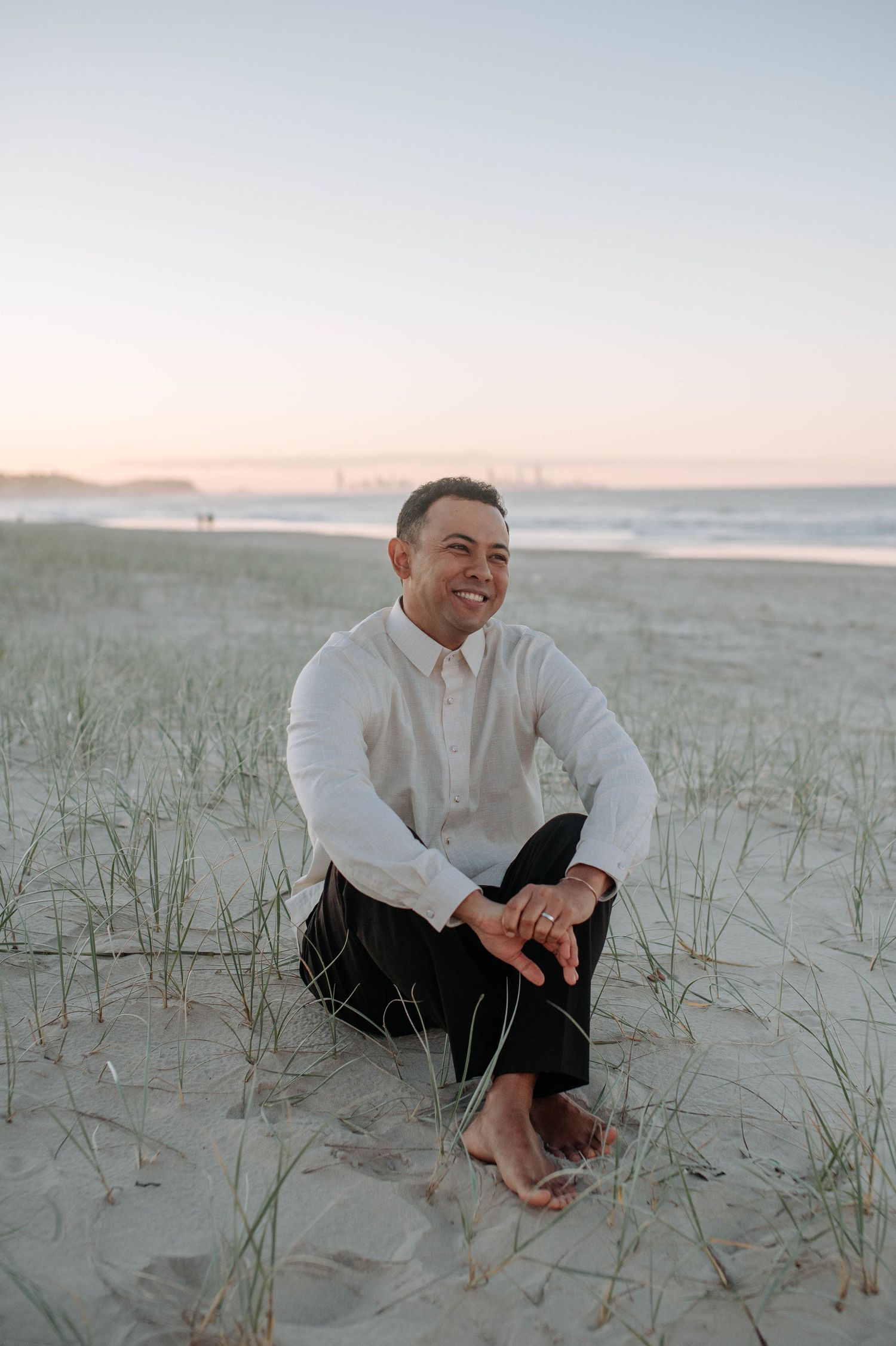 Someone sits smiling among beach grass at sunset wearing a white button-down shirt and dark pants with bare feet in the sand.
