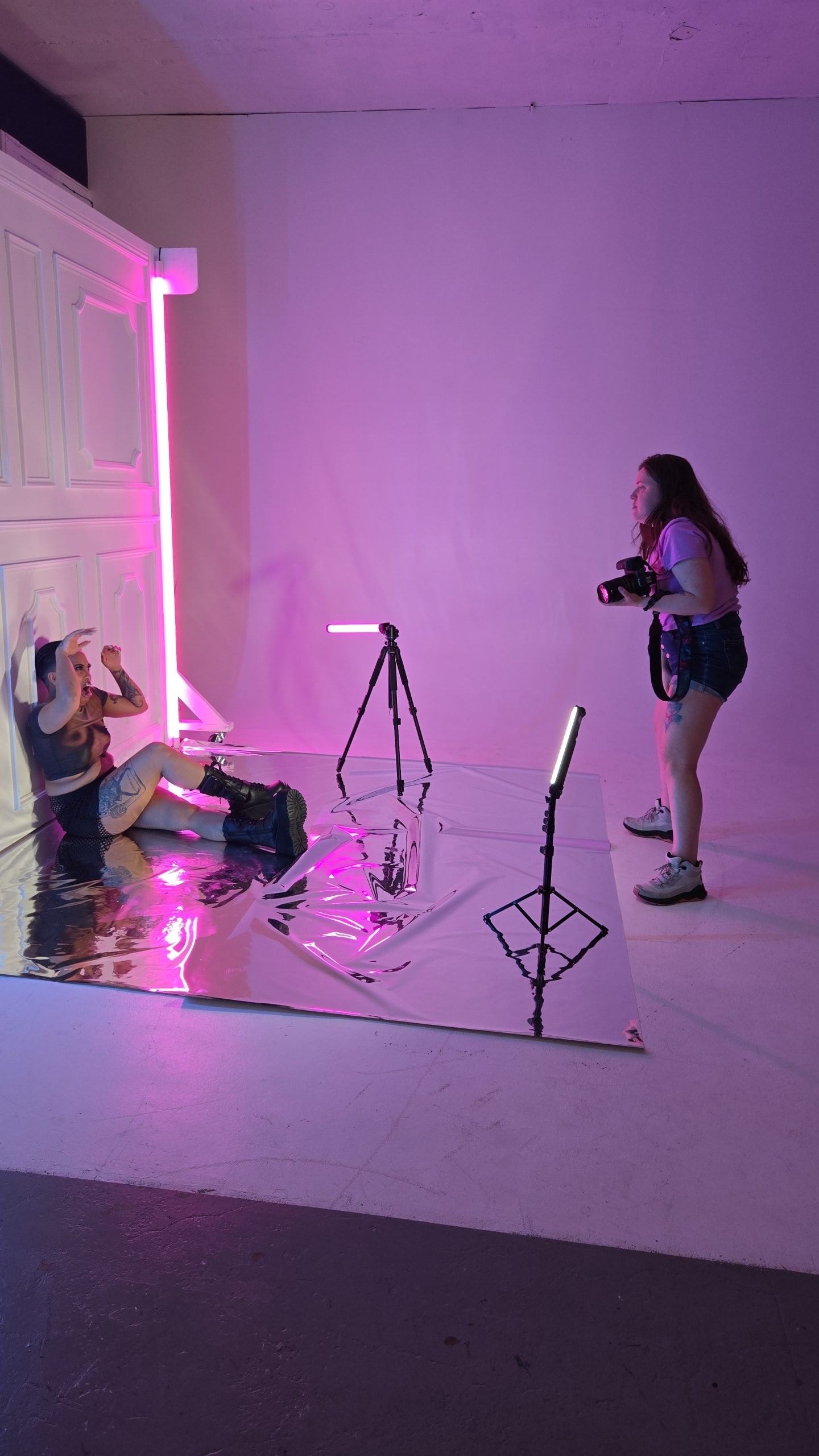 Behind-the-scenes photo shoot setup with pink neon lighting, camera equipment and tripods in a white studio space.