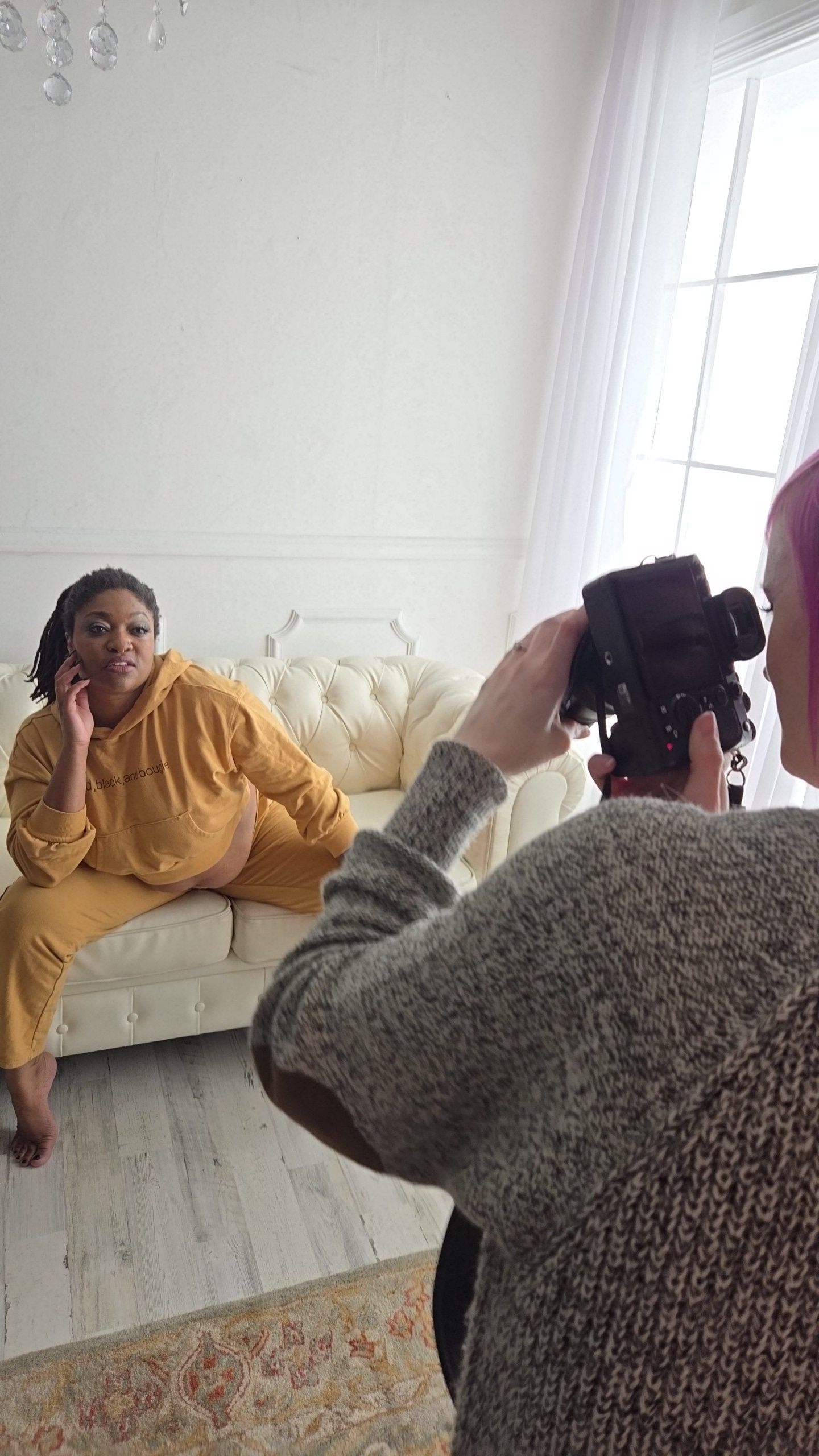 A photographer takes pictures of someone posing on a white tufted couch during a photo session in a bright studio.