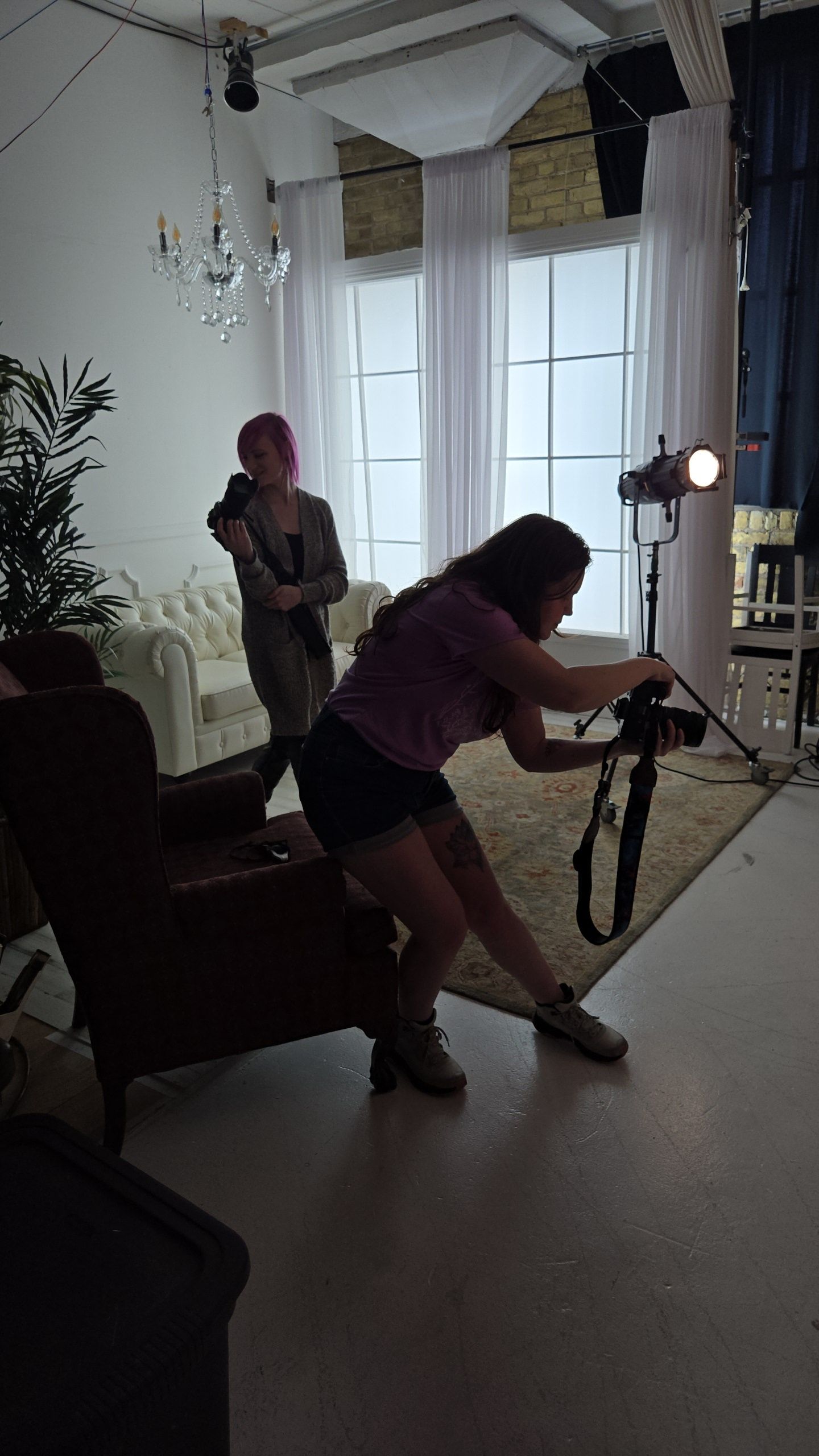Photographer with burgundy hair sets up equipment in a studio space with white curtains and chandelier lighting.
