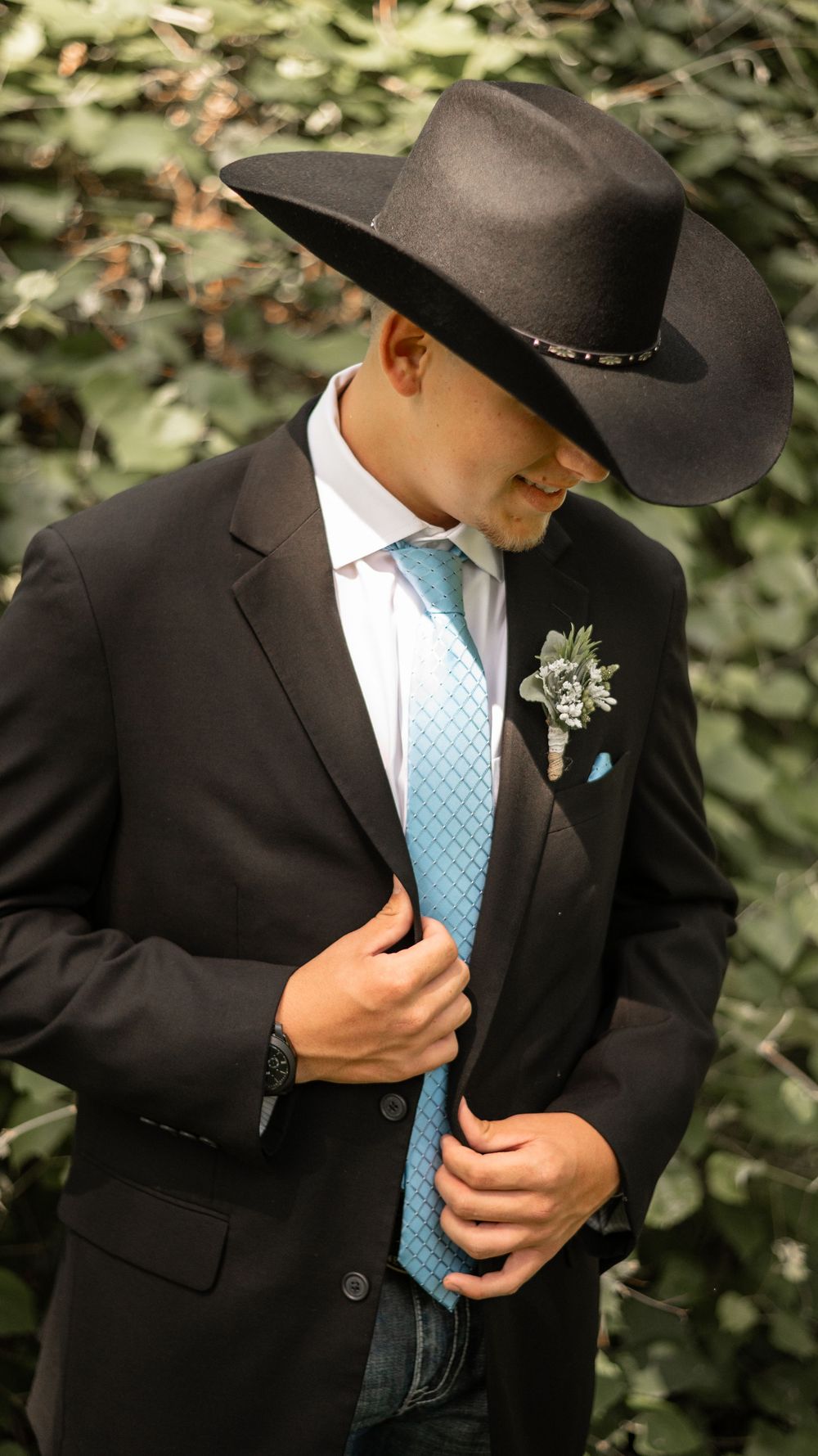 A wedding look featuring a black suit, light blue tie, and black cowboy hat with a boutonniere against a leafy backdrop.