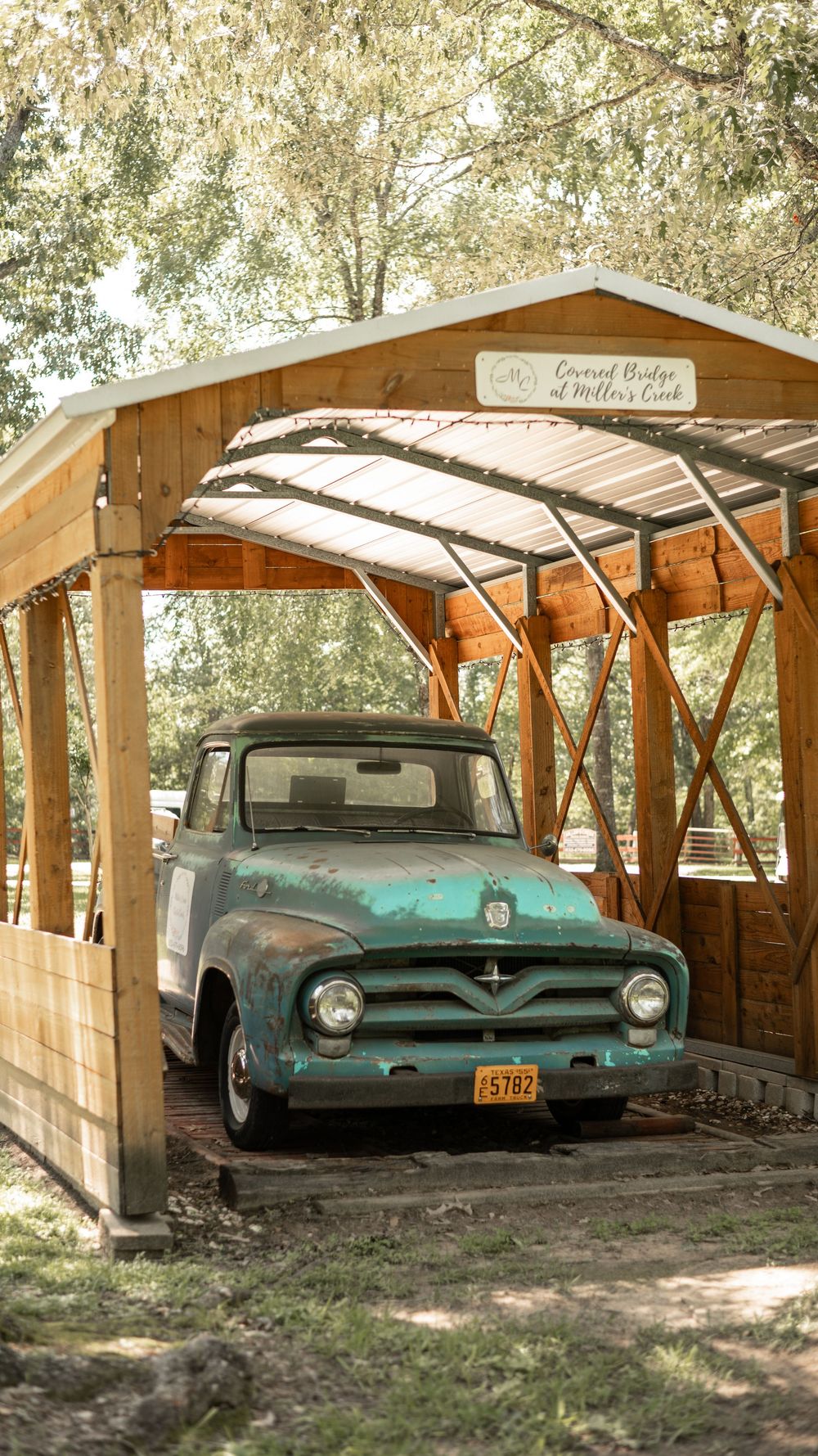 Vintage turquoise Ford pickup truck parked under a rustic wooden covered bridge structure.
