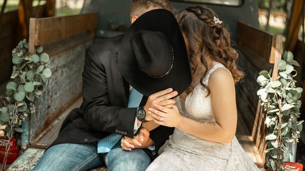 Couple holds hands in romantic wedding setting with floral decor and rustic wood backdrop in warm lighting.