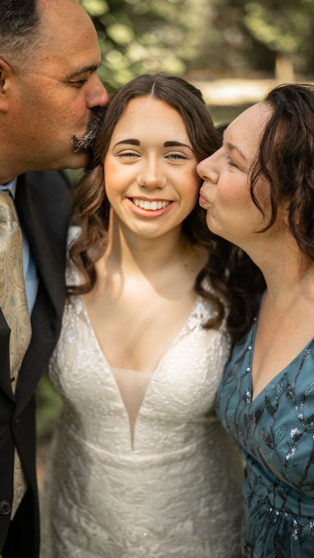 A happy bride in a white lace wedding dress shares a tender moment with family members showing genuine joy and affection.