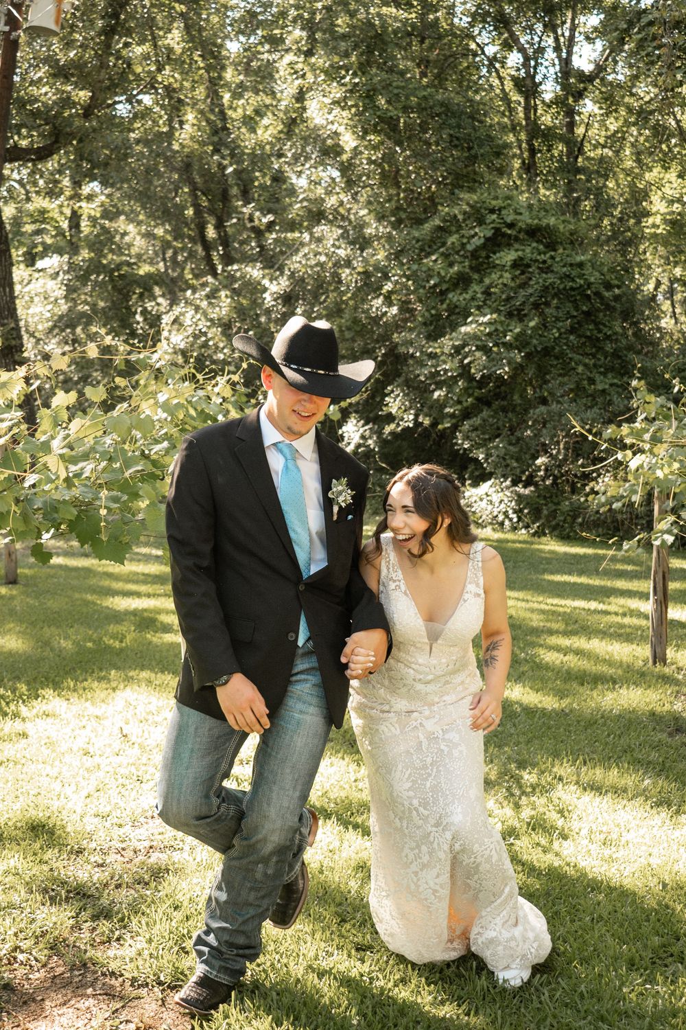 A couple walking together outdoors, with the groom in western attire and the bride wearing a lace wedding dress.