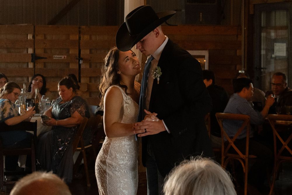 A couple shares their first dance at a rustic barn wedding reception with guests seated in the background.
