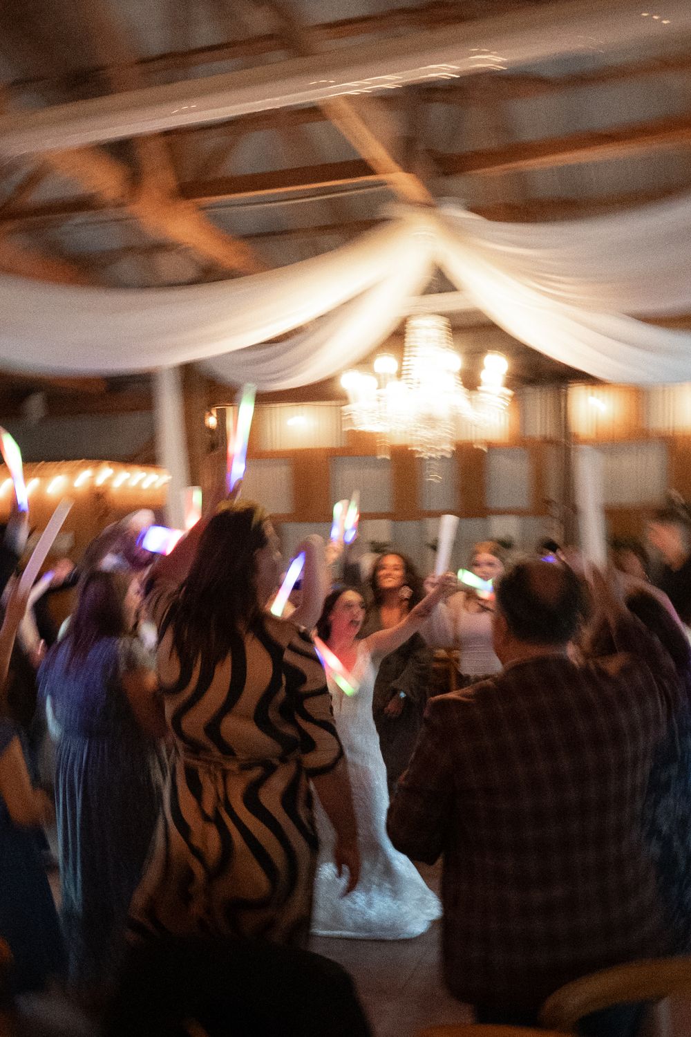Wedding guests dancing on a dimly lit dance floor under elegant white ceiling drapes and crystal chandeliers.