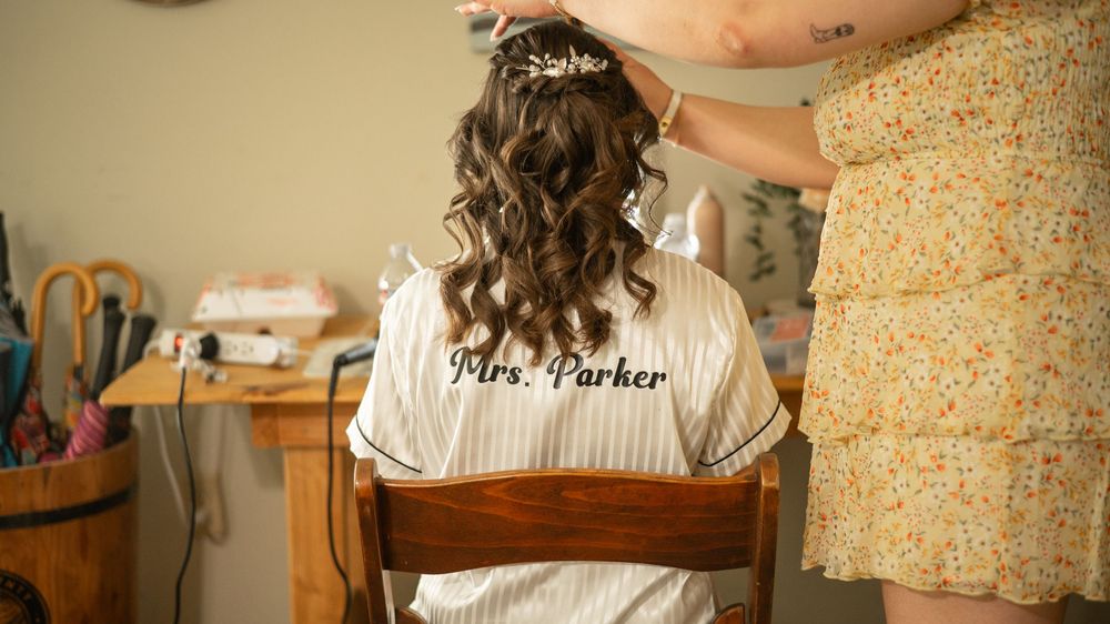 A bride sits in a chair getting her hair styled, wearing a white robe with brown sash against floral wallpaper.