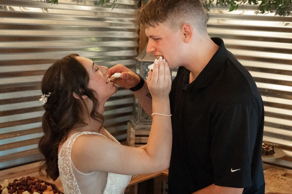 A couple playfully feeds each other wedding cake at their reception in front of a corrugated metal backdrop.