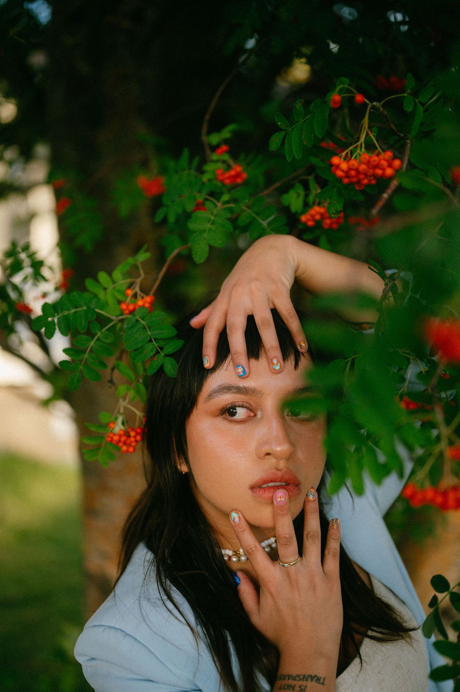 Artistic portrait beneath red rowan berries and green leaves in natural outdoor lighting with moody autumn aesthetic.