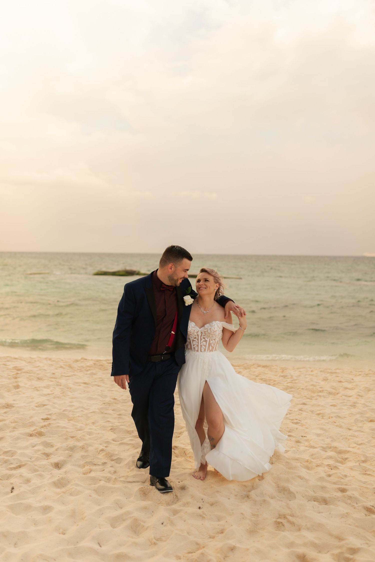 Bride and groom walking on sandy beach at sunset with ocean in background.