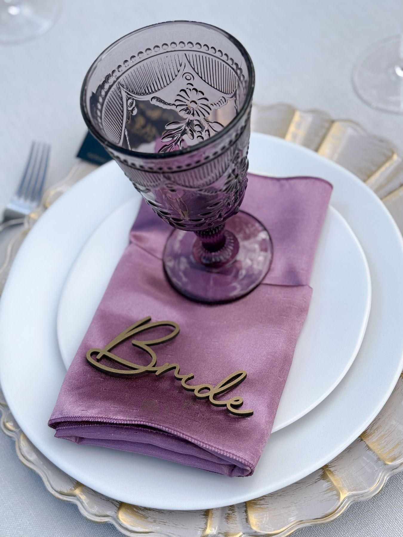 Elegant place setting with purple vintage glass goblet, white plate, and lavender napkin with gold 'Bride' text.