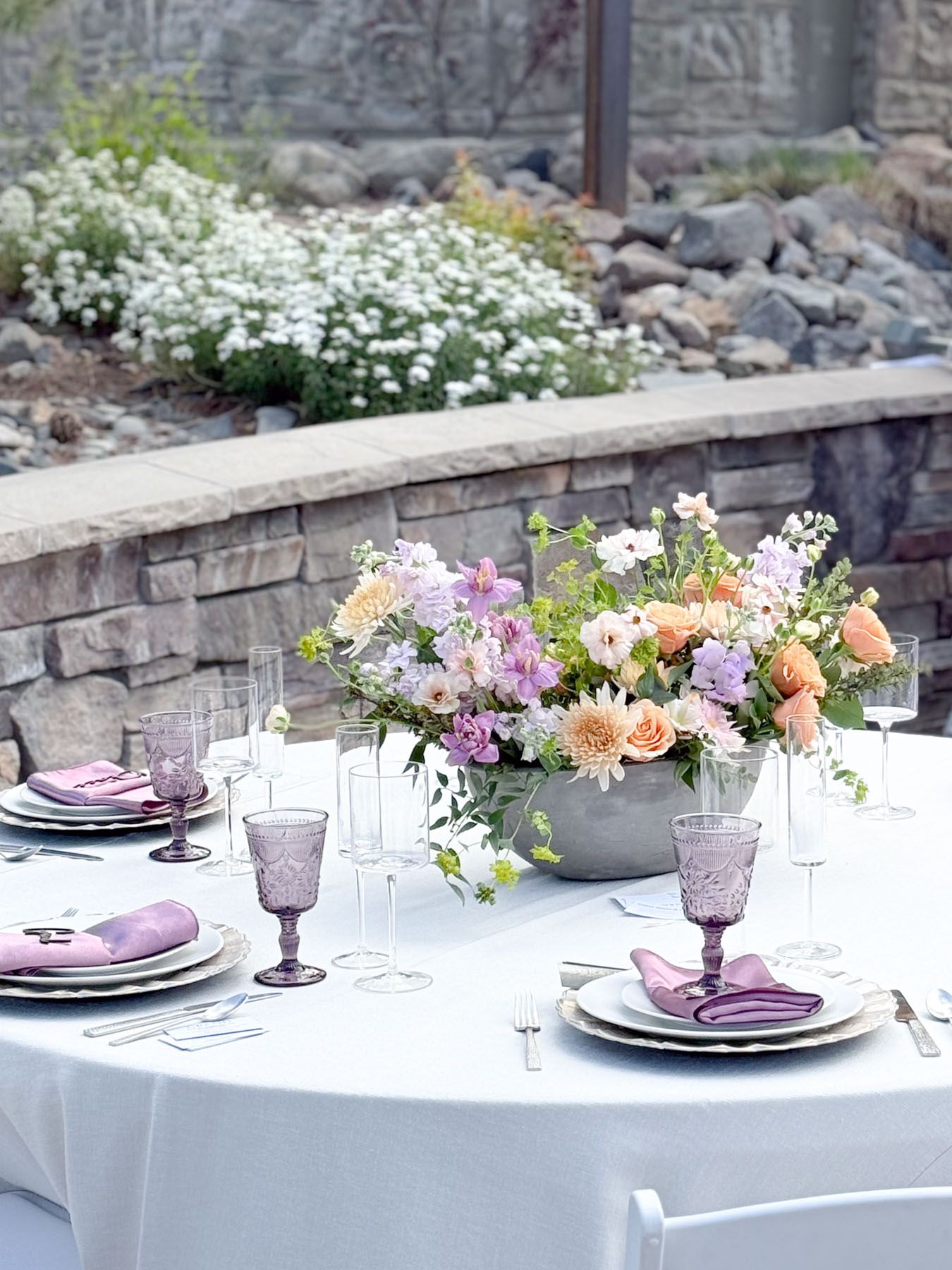 An elegant outdoor table setting with purple glasses, white plates, and a centerpiece of peach and pink flowers in a stone planter.