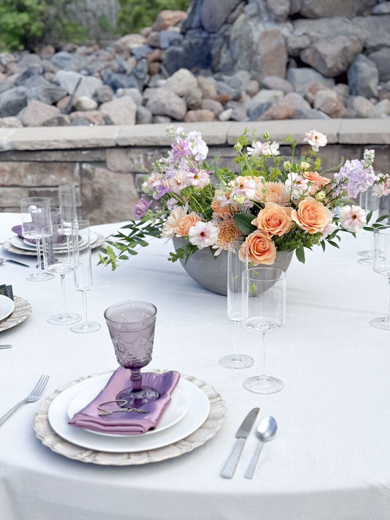 Elegant table setting with peach and purple flowers, purple glassware, and white dishes against a rocky outdoor backdrop.