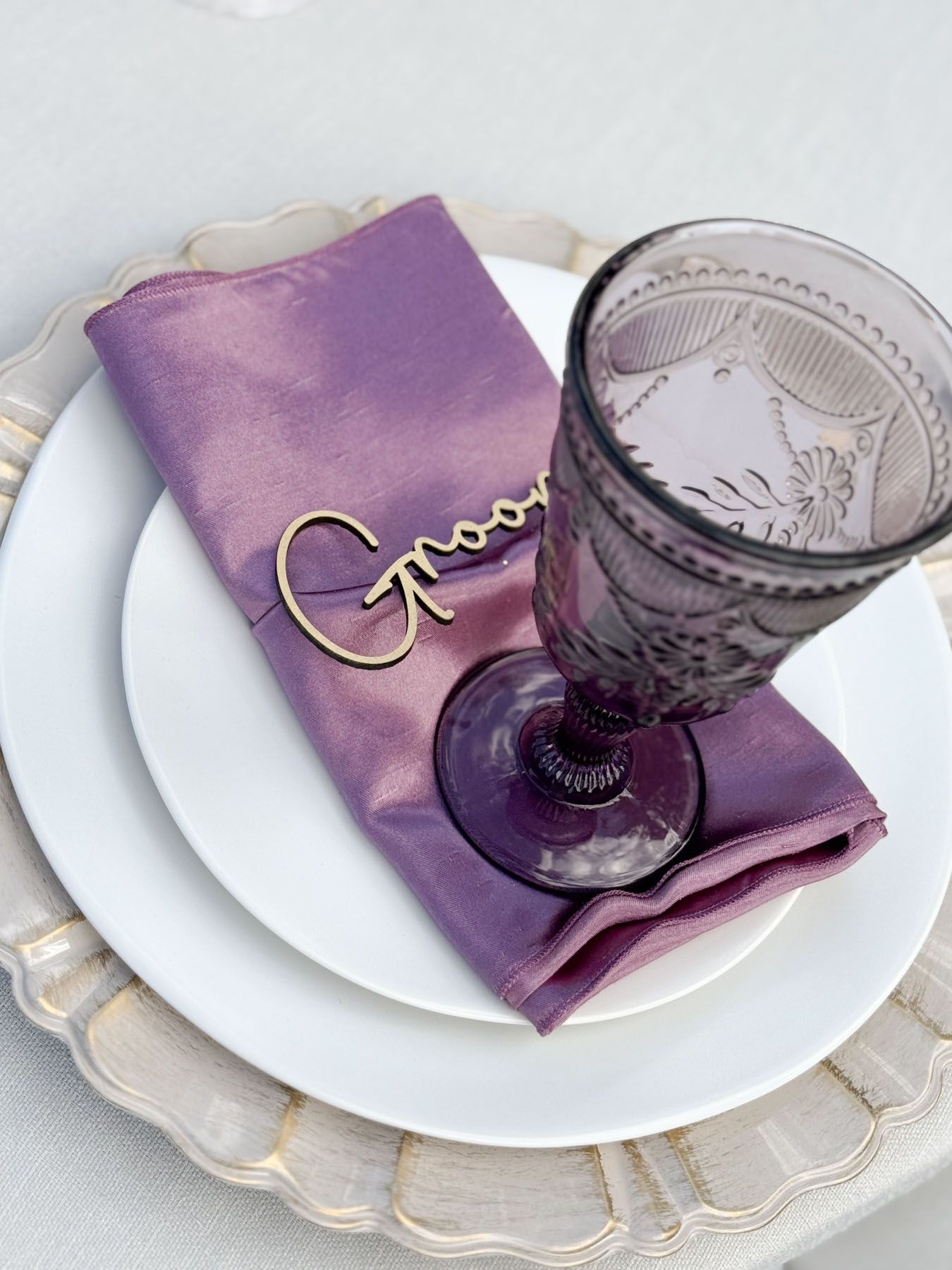 An elegant place setting with purple napkin, decorative name tag, and amethyst-colored glass on a scalloped charger plate.