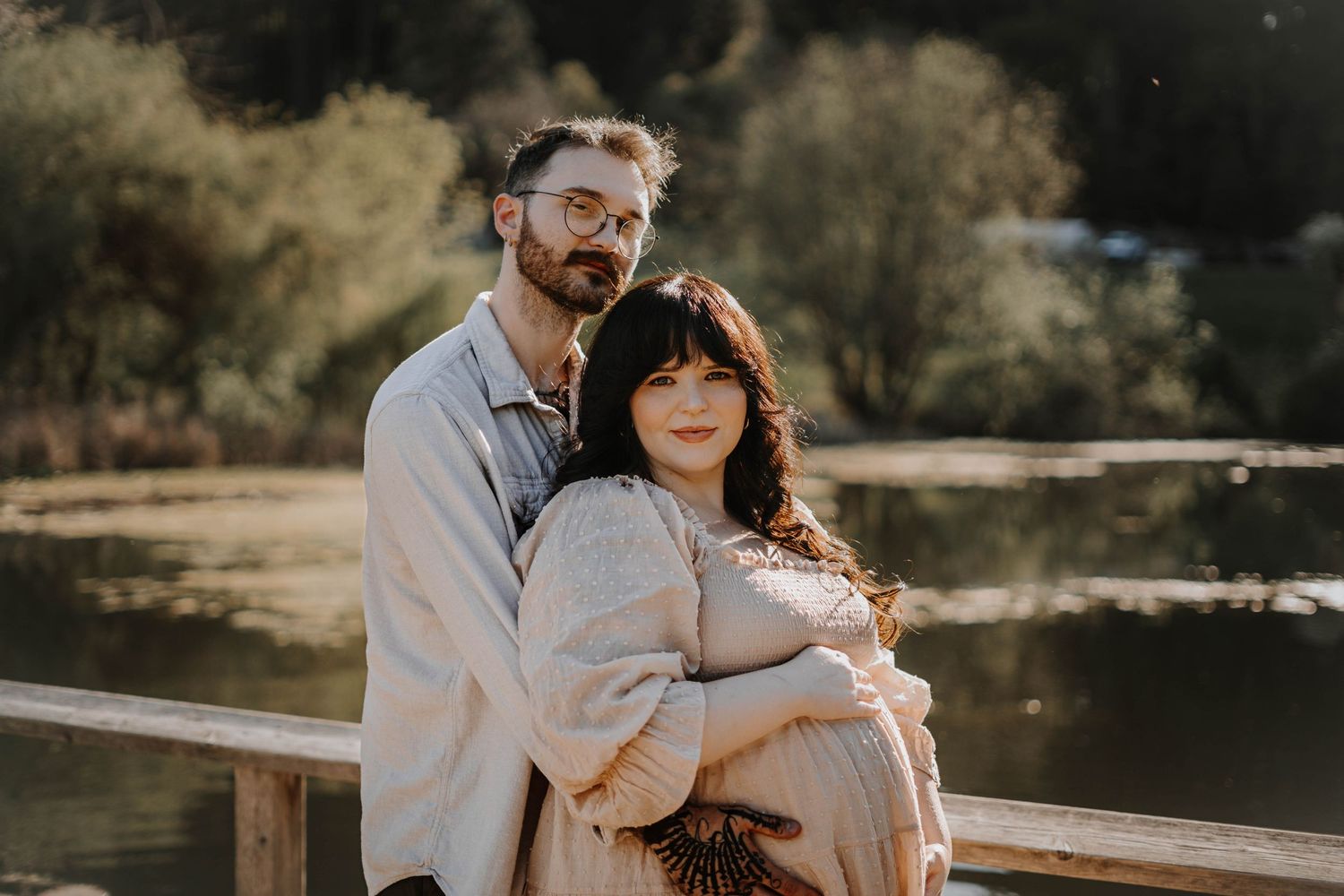 Maternity couple portrait with pregnant woman in cream dress embraced by bearded man near serene lake.