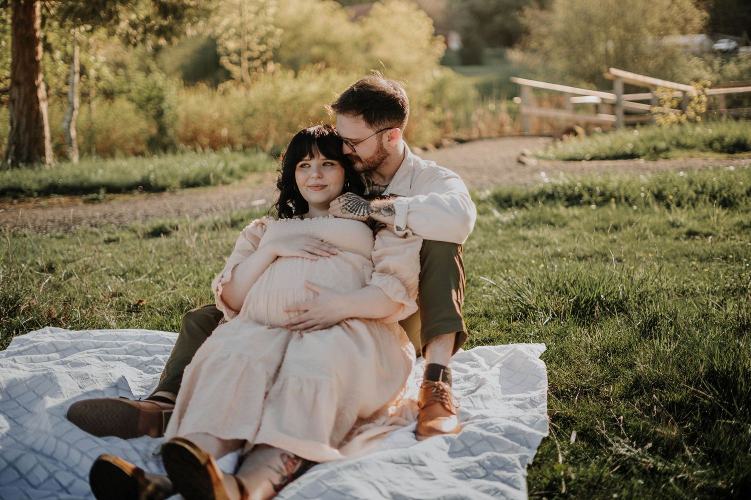 Couple sharing intimate maternity photo on white blanket in golden sunlit outdoor setting.
