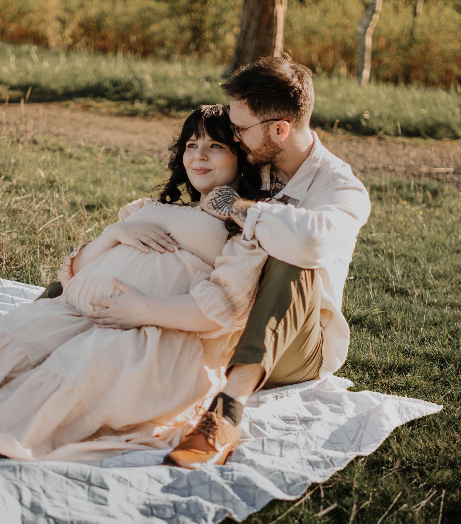 Pregnant woman in blush maternity gown sitting on blanket with partner in sunny green meadow.