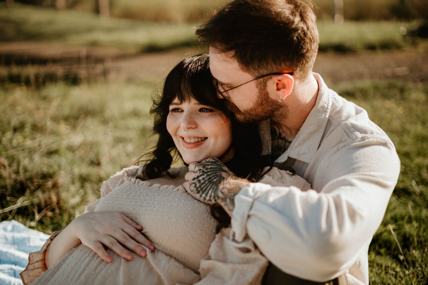 Smiling pregnant woman in cream dress cuddled by bearded partner with glasses on grass blanket.