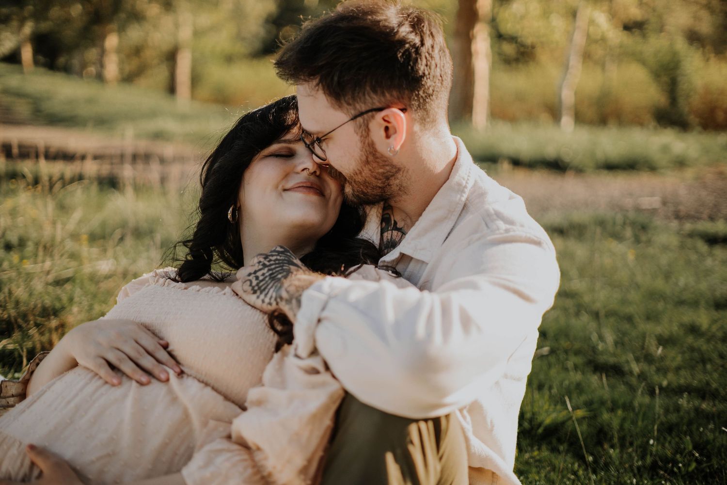Couple sharing a tender kiss during outdoor maternity session on a blanket in a green field.