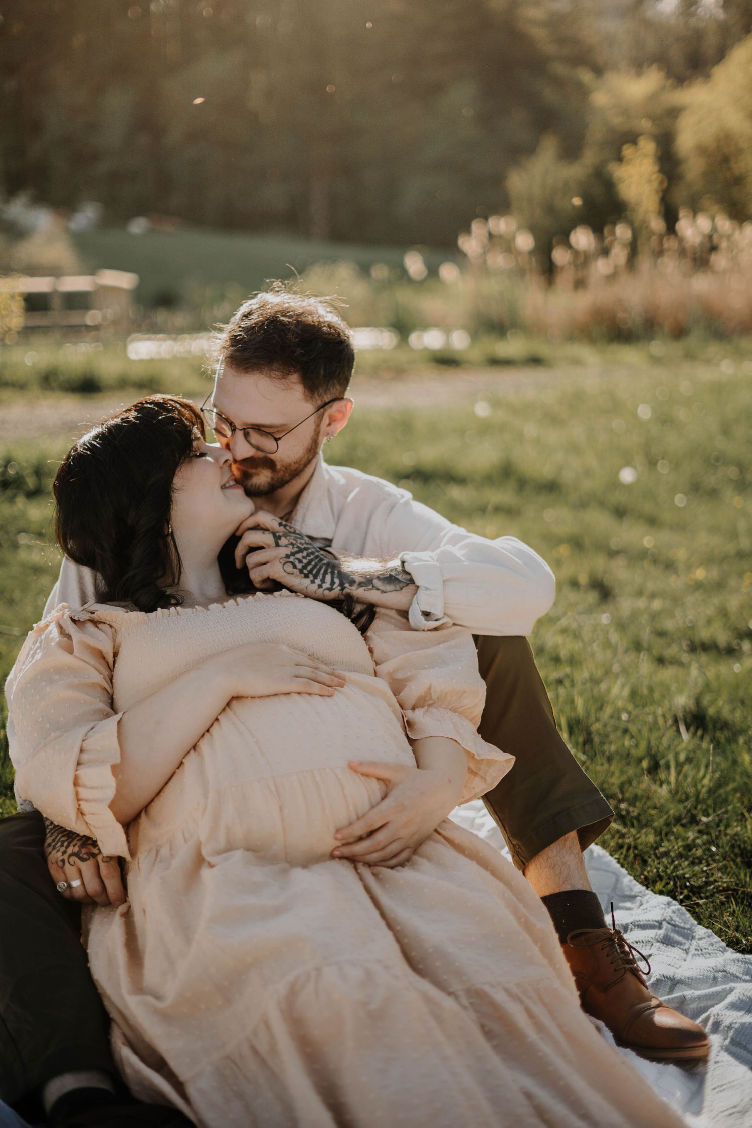 Couple sharing a tender kiss during golden hour maternity photoshoot on a blanket in a lush green field.