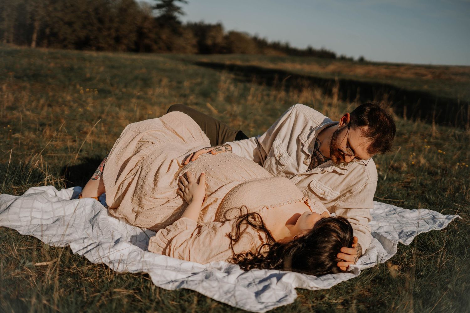 Pregnant couple lying on a white blanket in a golden field, sharing an intimate and joyful moment.