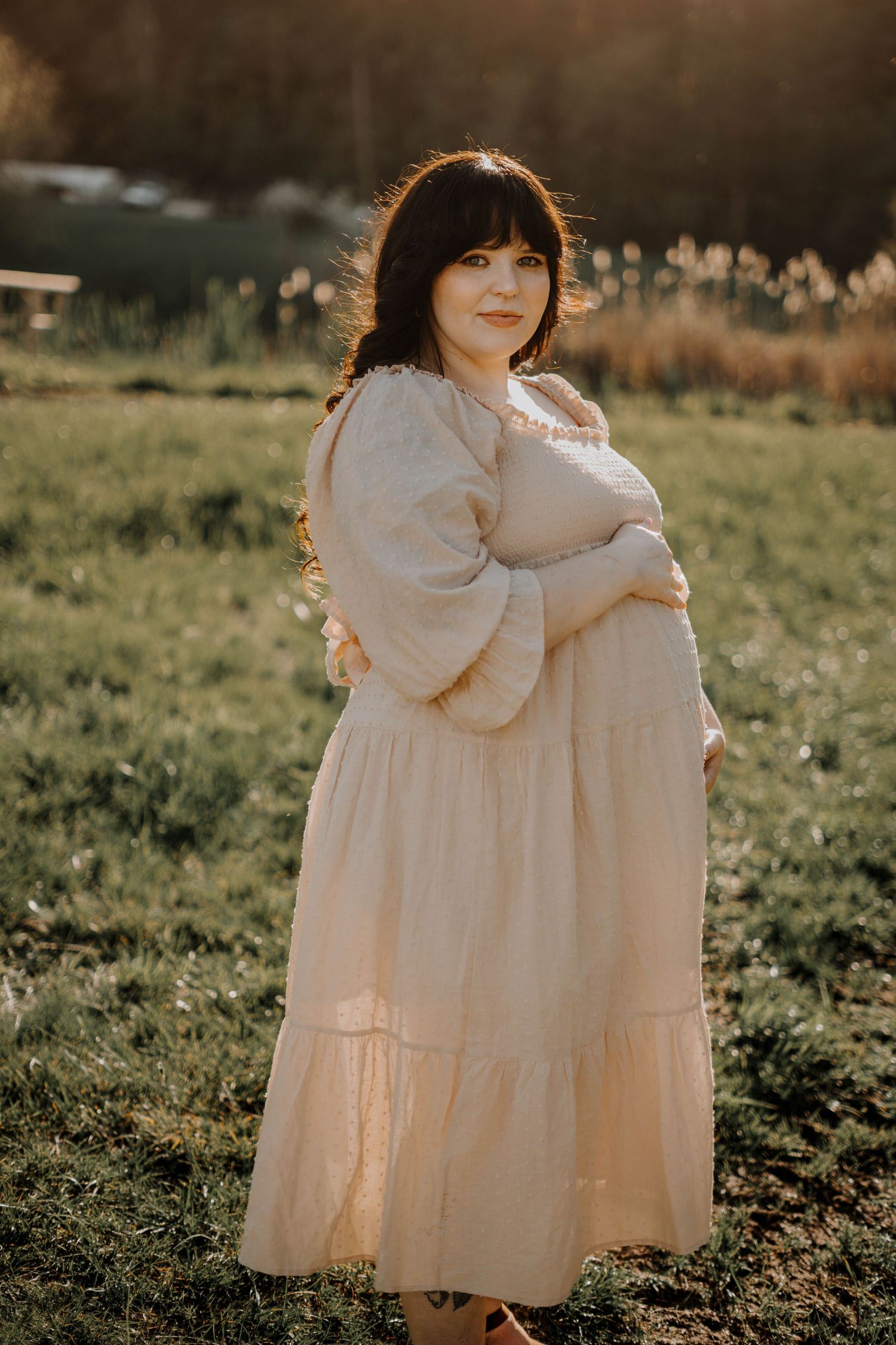 Pregnant woman in a flowing cream boho dress cradling her baby bump during a golden hour outdoor shoot.