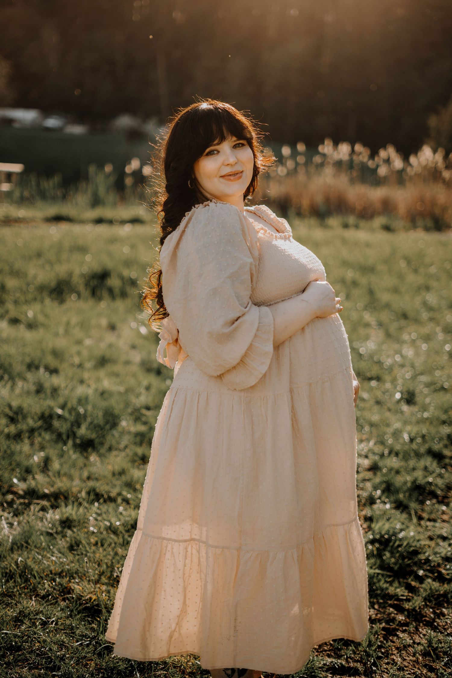 Smiling pregnant woman in a cream tiered maxi dress posing in a sunlit green field at sunset.