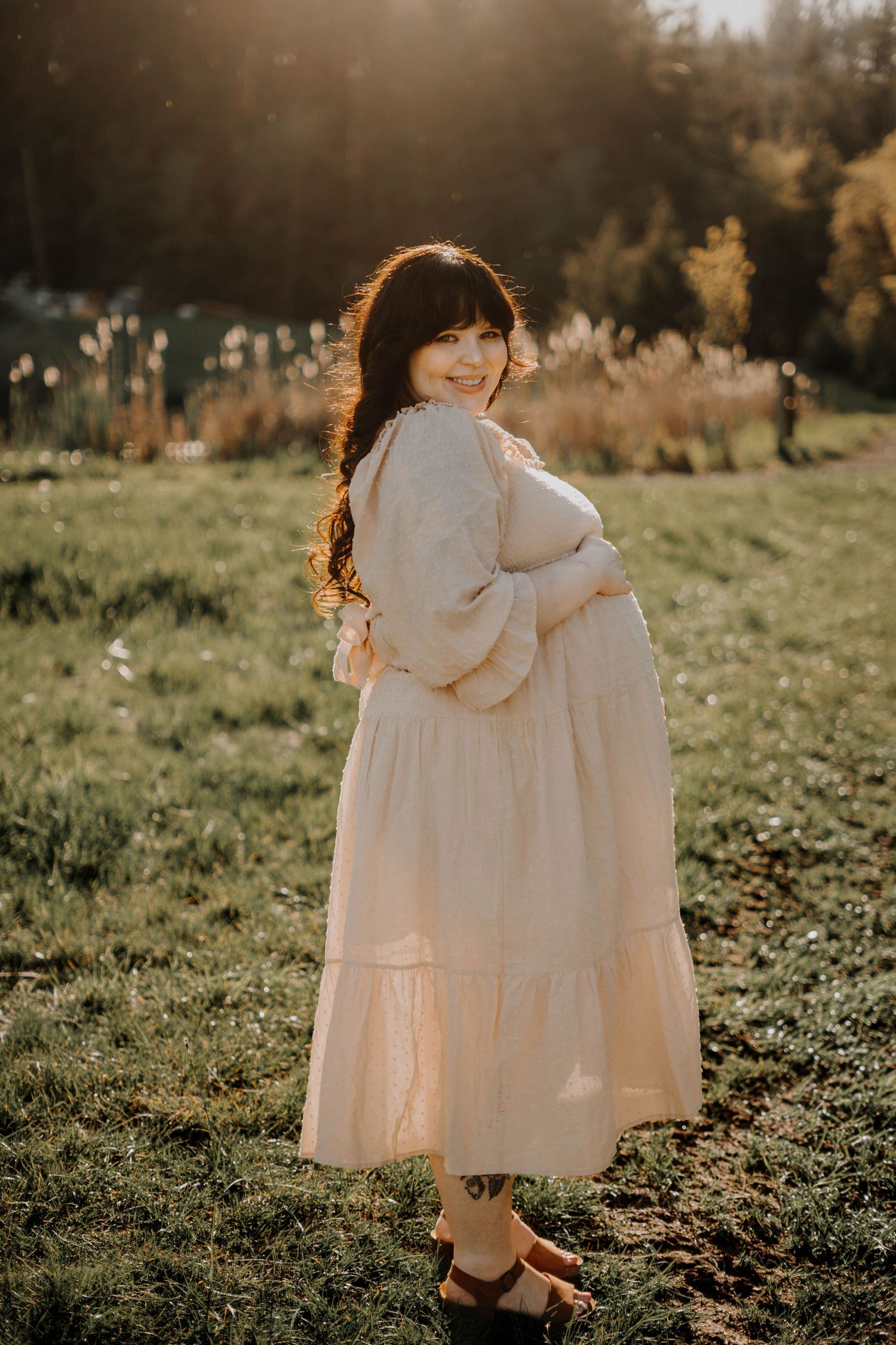 Pregnant woman in a cream boho dress smiling over her shoulder in a glowing golden hour field setting.