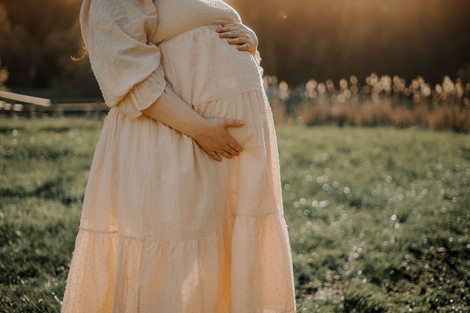 Close-up of a pregnant woman in a cream dress gently cradling her baby bump in a sun-drenched field.