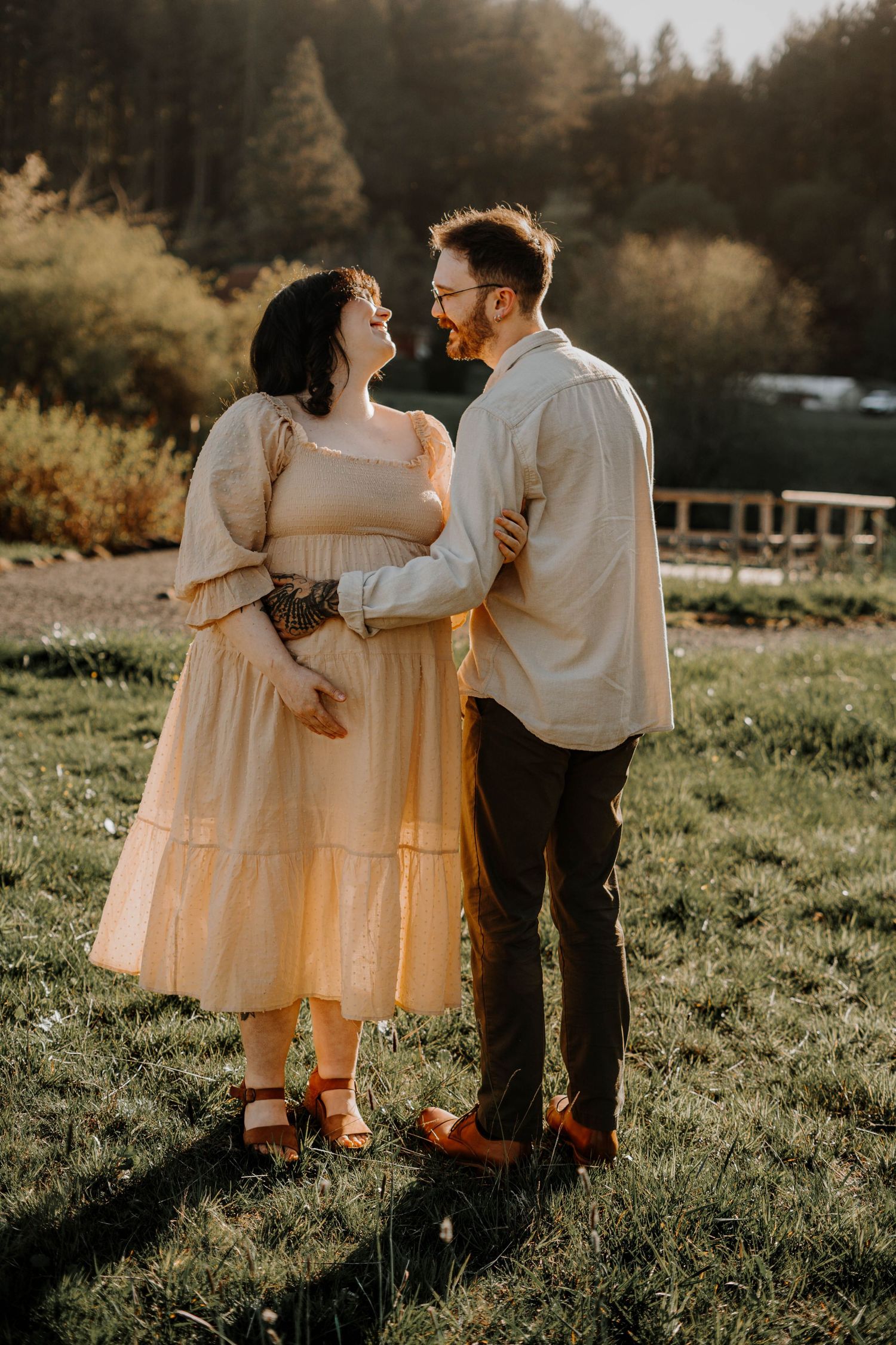 Expecting couple smiling at each other while embracing in a sunlit green field during maternity session.