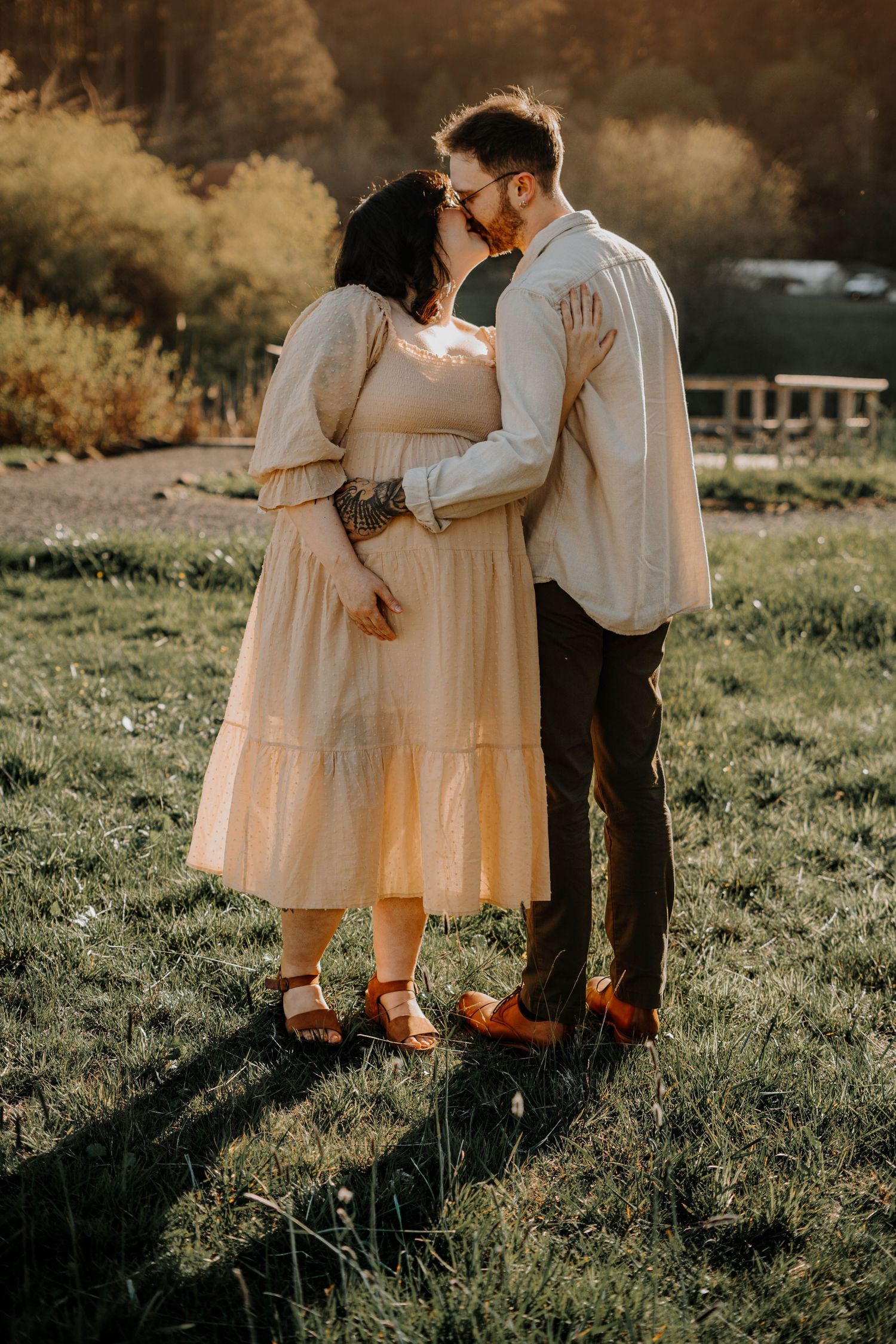 Couple sharing a romantic kiss during a golden hour maternity photoshoot in a scenic outdoor setting.