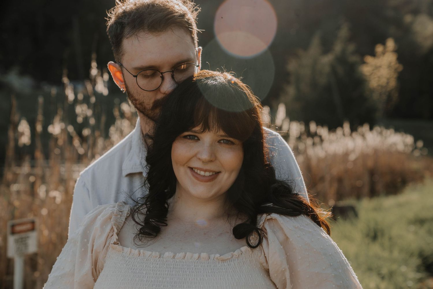 Couple smiling outdoors in golden sunlight, woman with dark hair in cream dress, man with glasses embracing her from behind.