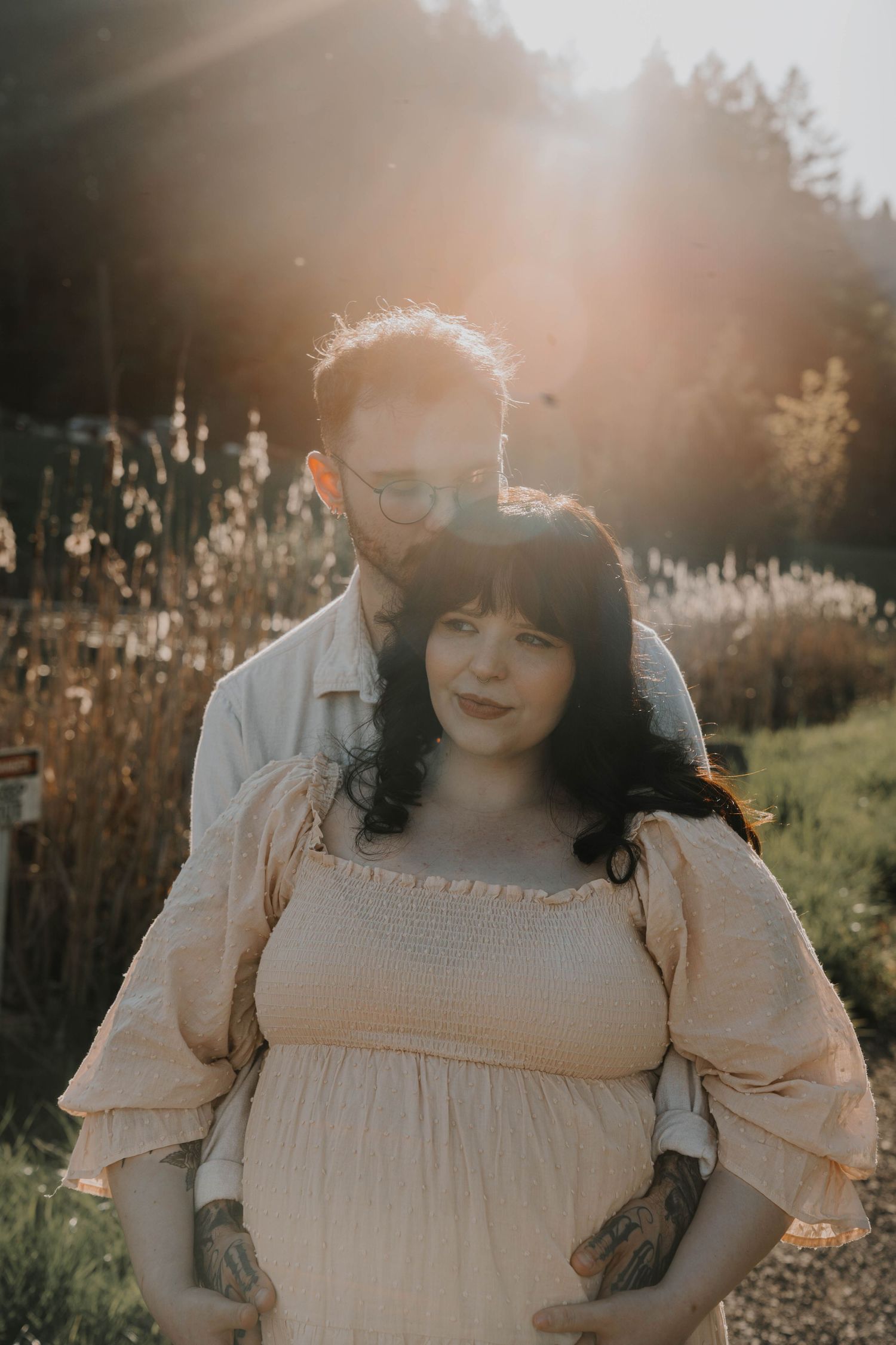 Pregnant woman in cream smocked dress with partner embracing her from behind in a sun-drenched garden setting.