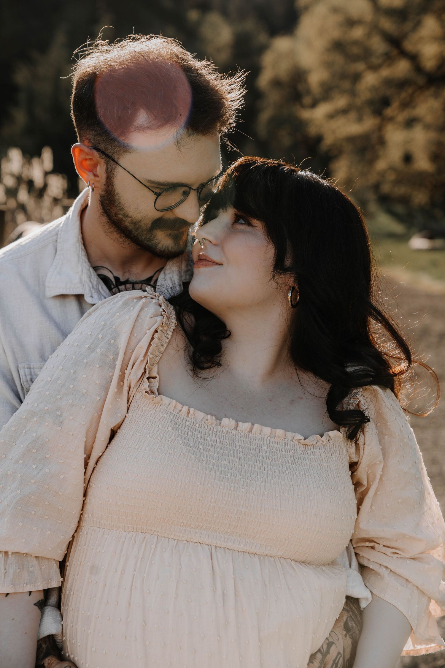 Couple sharing a tender moment outdoors, woman in cream off-shoulder dress leaning into man with glasses in sunlight.