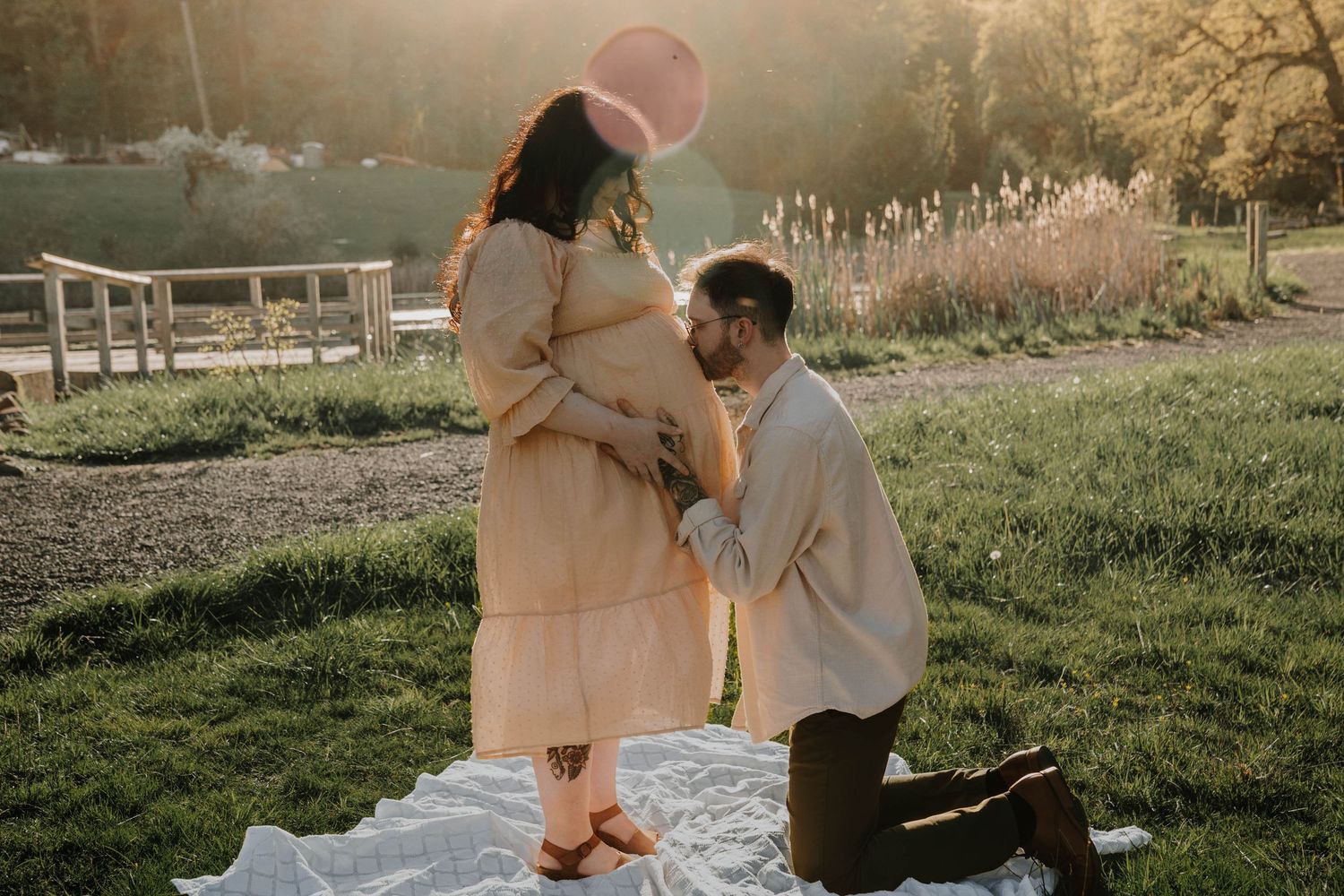 Man kneeling before pregnant woman holding pink balloon in a sunny garden, touching her baby bump tenderly.