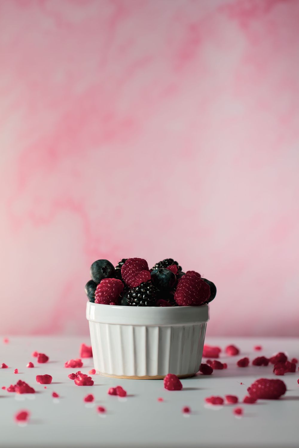 Fresh raspberries, blueberries and blackberries in a white ceramic ramekin against a pink background with scattered raspberry crumbs.