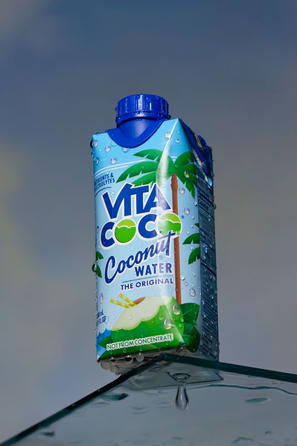 A carton of Vita Coco coconut water with palm tree graphics sitting on clear glass surface against blue sky background.