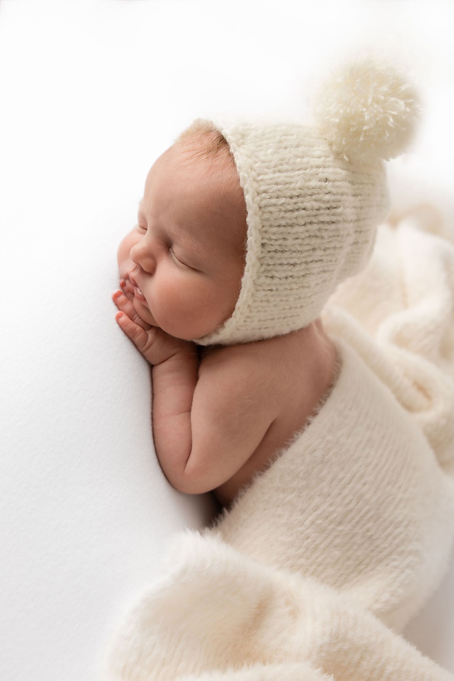 A sleeping newborn in a white knit bonnet and wrap rests peacefully against a soft white background.
