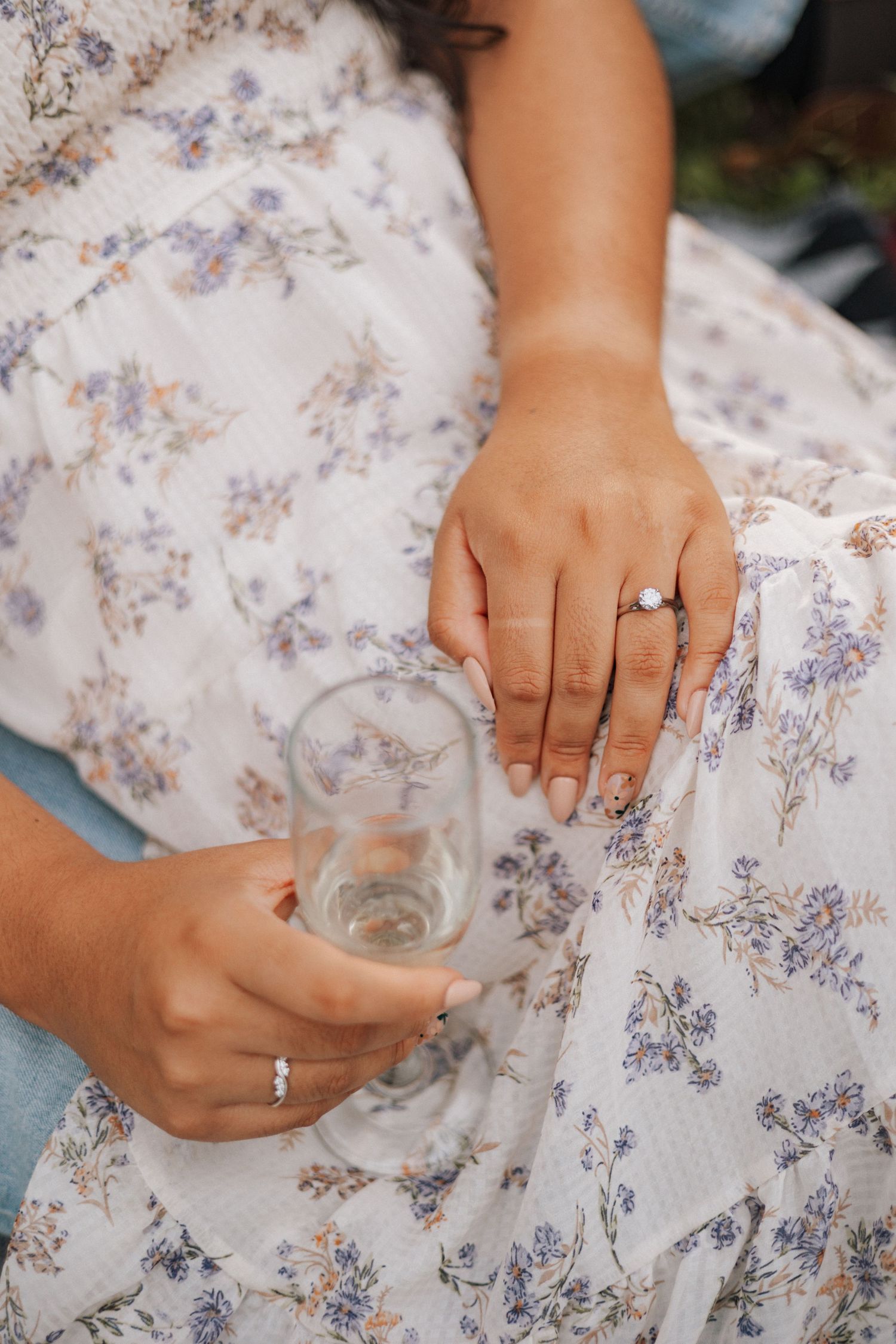 Hands in a floral dress hold a glass while displaying an engagement ring with a clear stone.