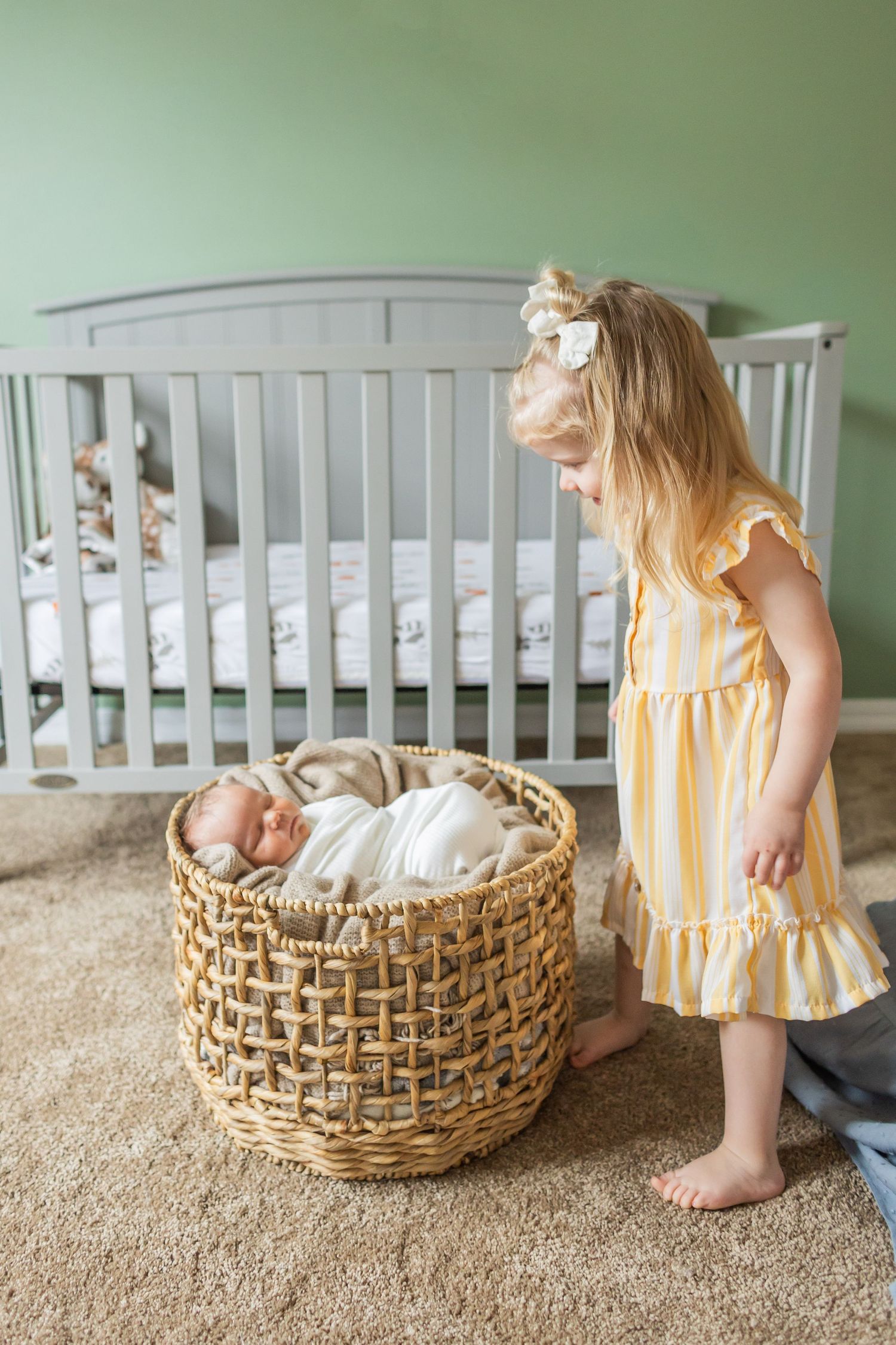 A young child in a yellow dress stands next to a woven basket containing a sleeping baby in a nursery with a grey crib.