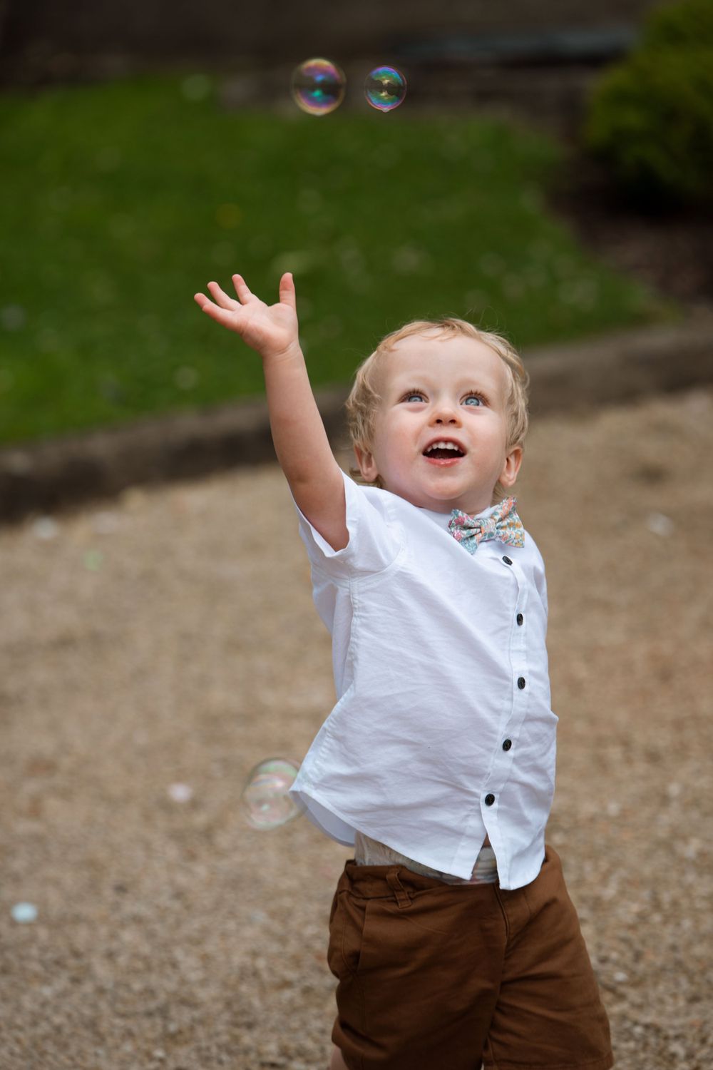 Young child in white shirt and brown pants reaches for bubbles during outdoor celebration.