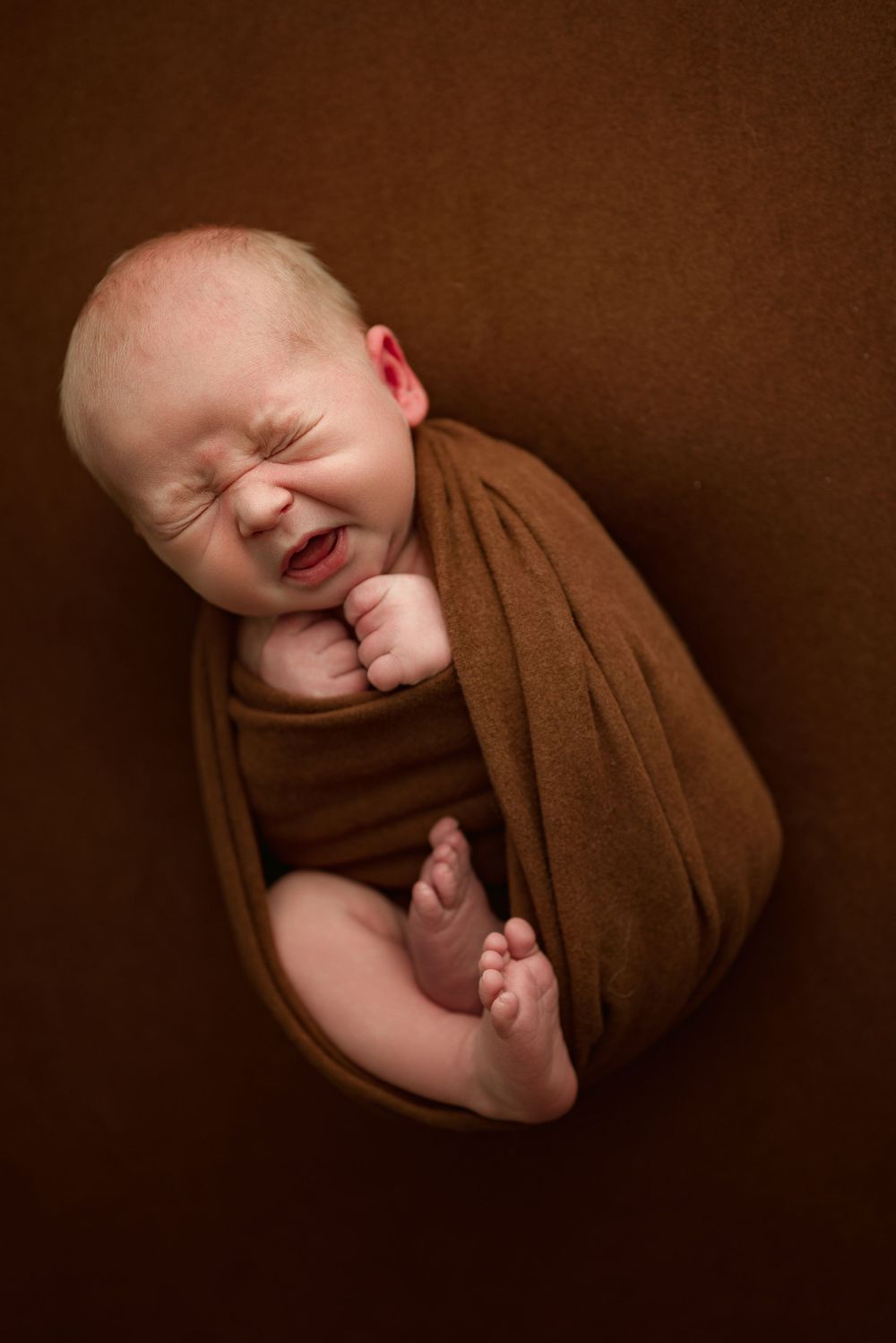 Newborn baby boy yawns and stretches while wrapped in brown swaddle during studio newborn session in Clarksville, Tennessee.