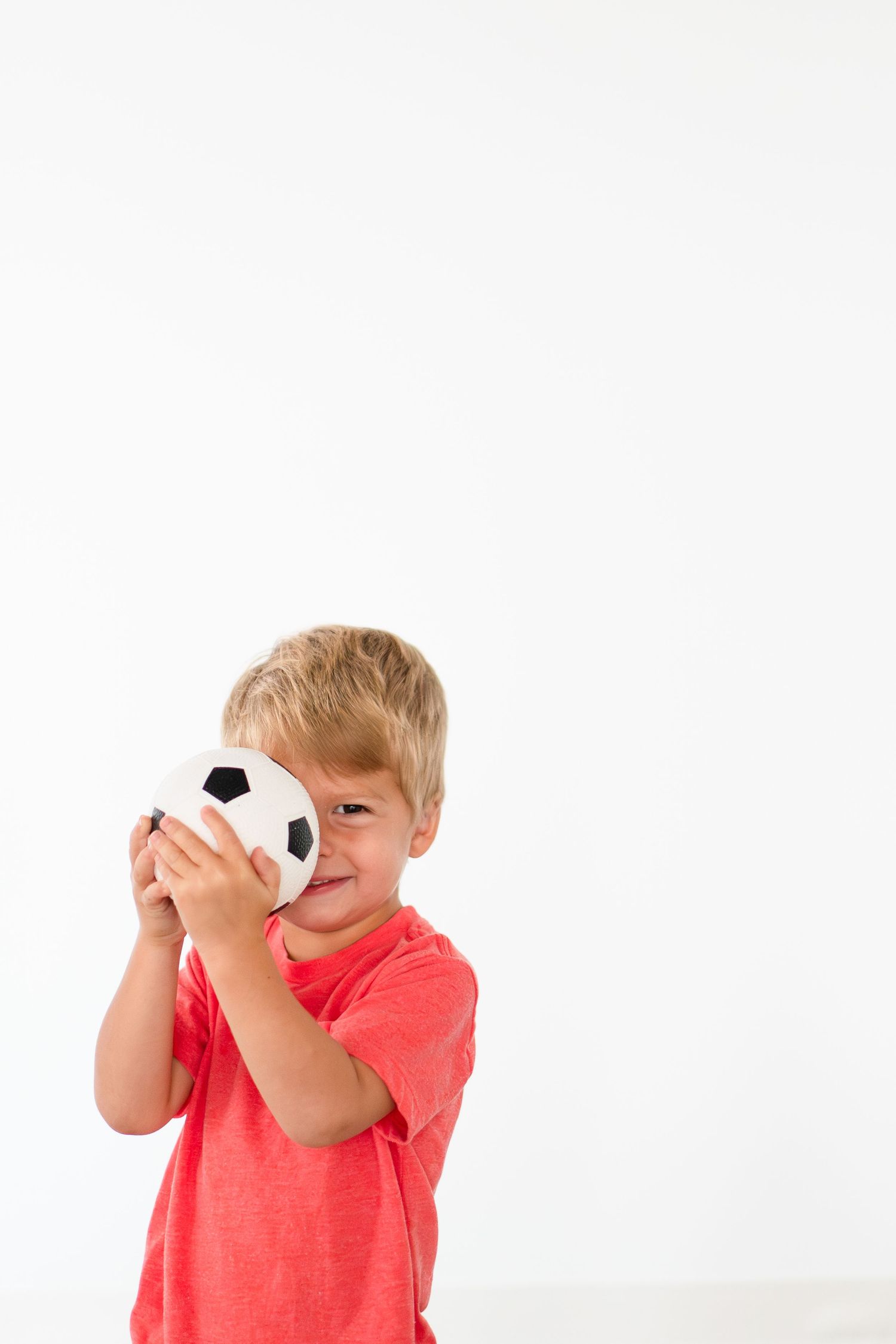 Young child in coral t-shirt holds small soccer ball plush toy against white background.