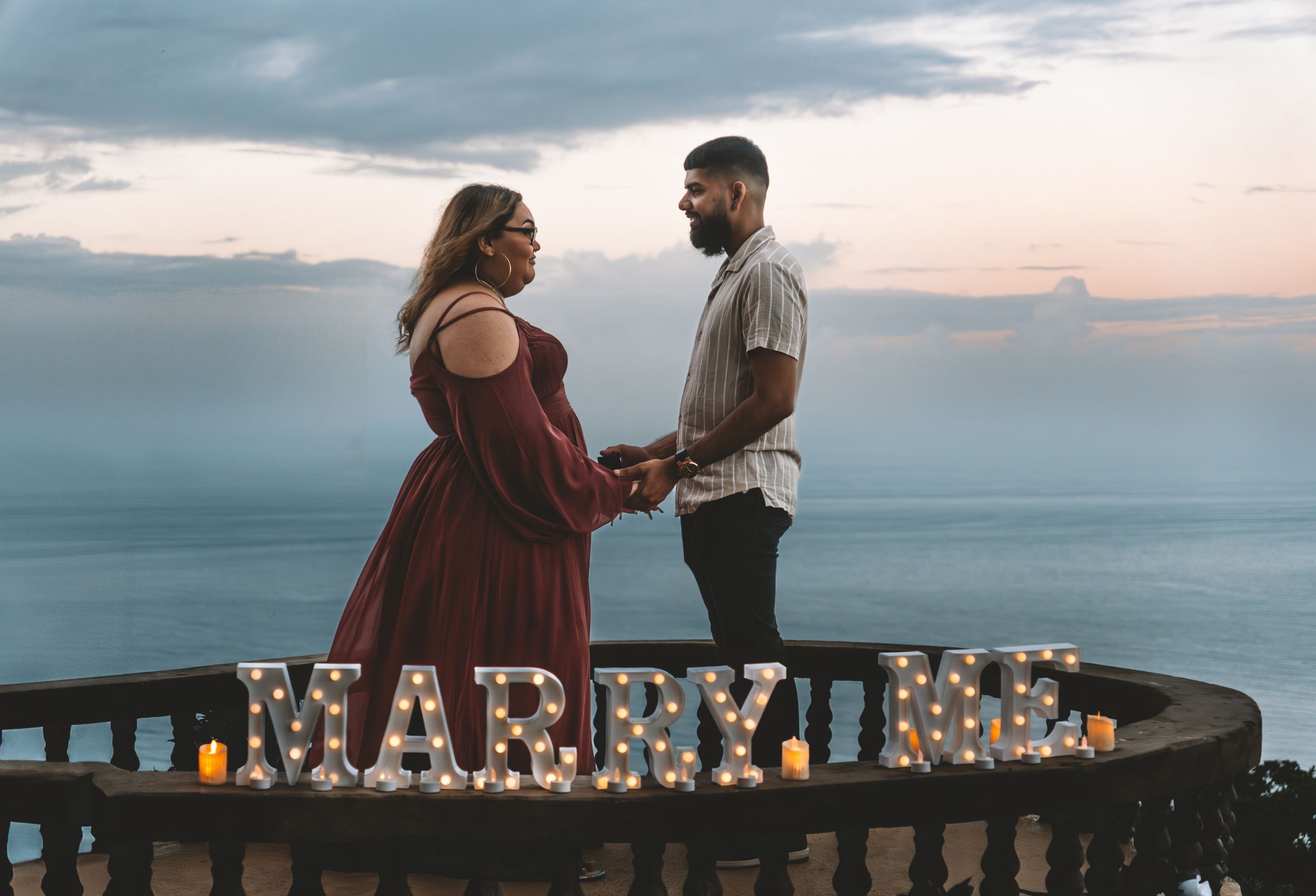 Romantic couple standing on a balcony at sunset with illuminated 'MARRY ME' lettering in the background.